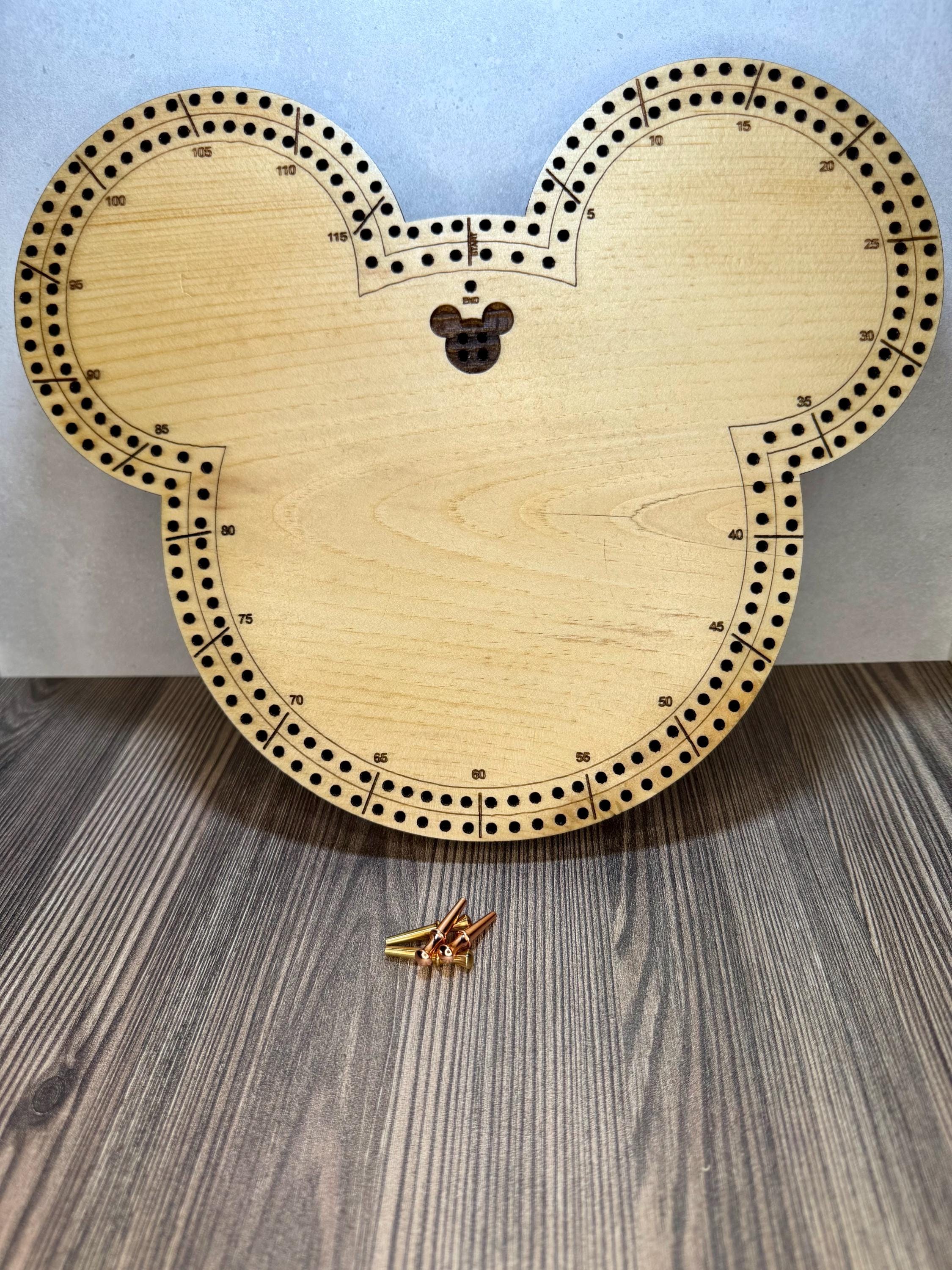 Handmade wooden mouse-shaped cribbage board standing upright on a wood surface, with engraved scoring tracks and metal pegs placed in front for scale.