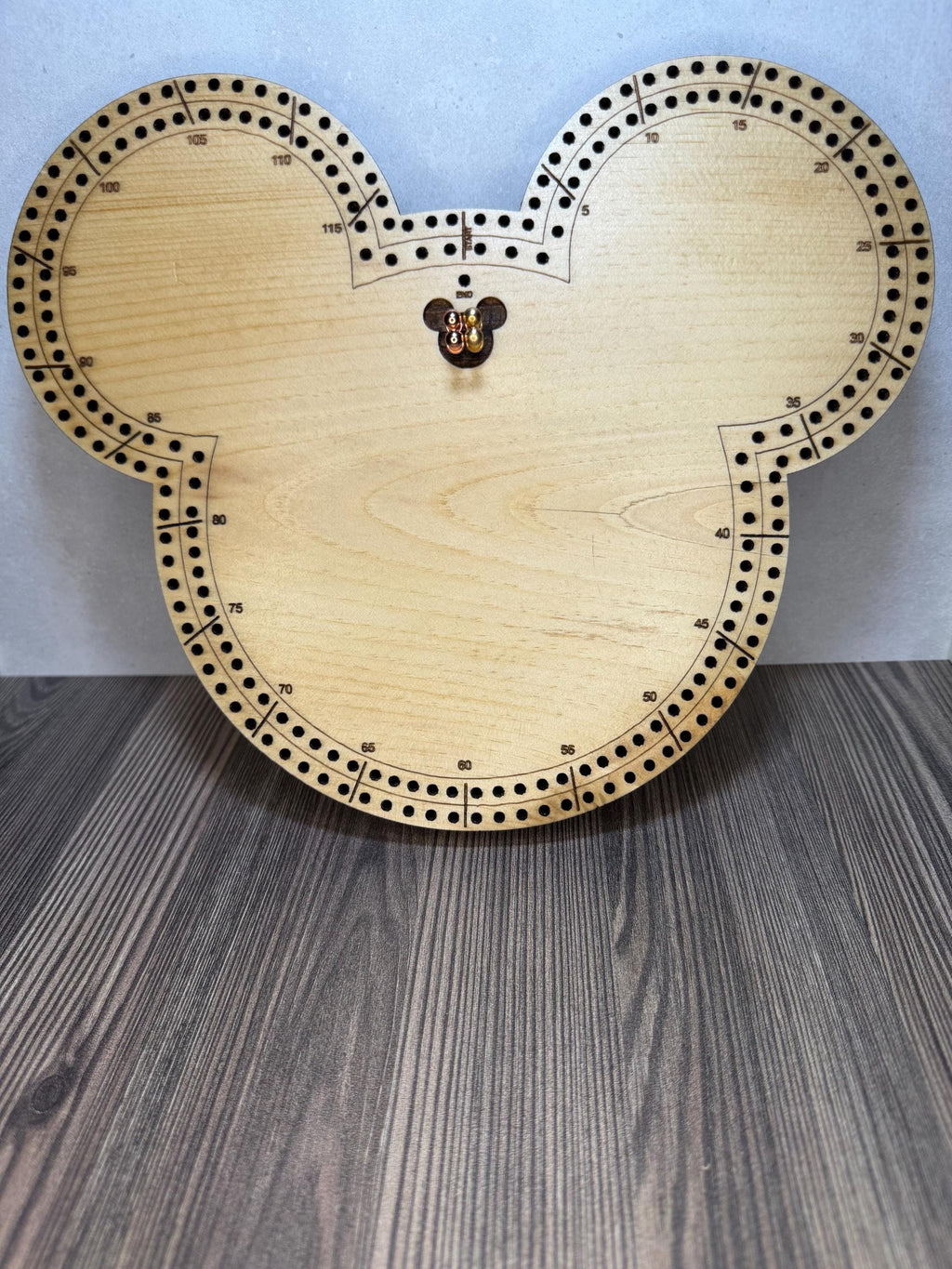 Front view of a handmade wooden mouse-shaped cribbage board standing upright on a wood surface, featuring engraved scoring tracks with metal pegs placed in the center.