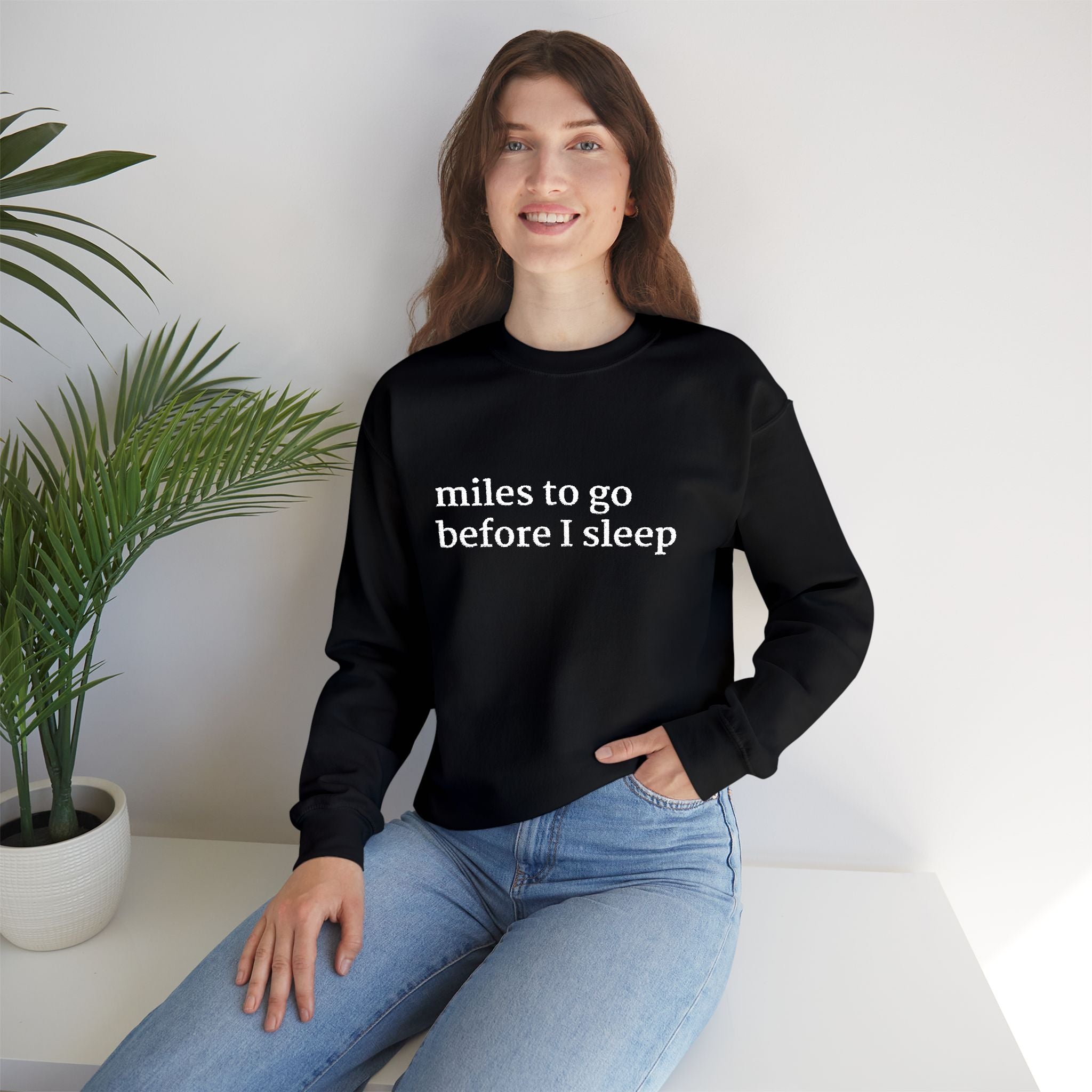 Model wearing a black crewneck sweatshirt with white text reading ‘miles to go before I sleep,’ paired with light blue jeans.