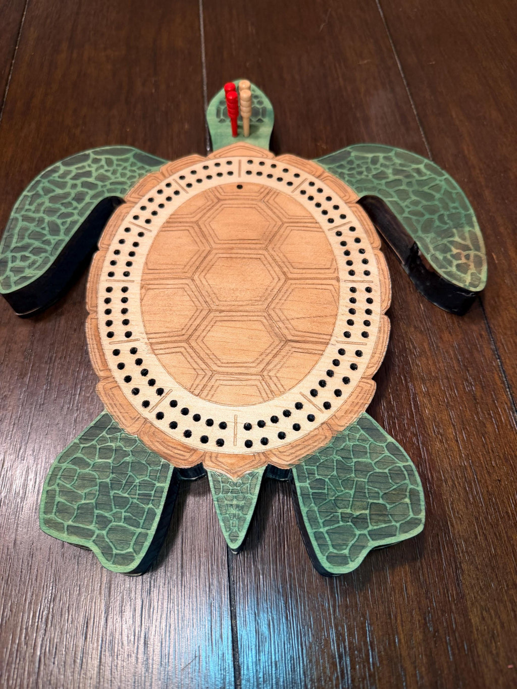 Overhead view of a wooden turtle cribbage board with four cribbage pegs placed at the starting area, showing the full board layout and engraved details.