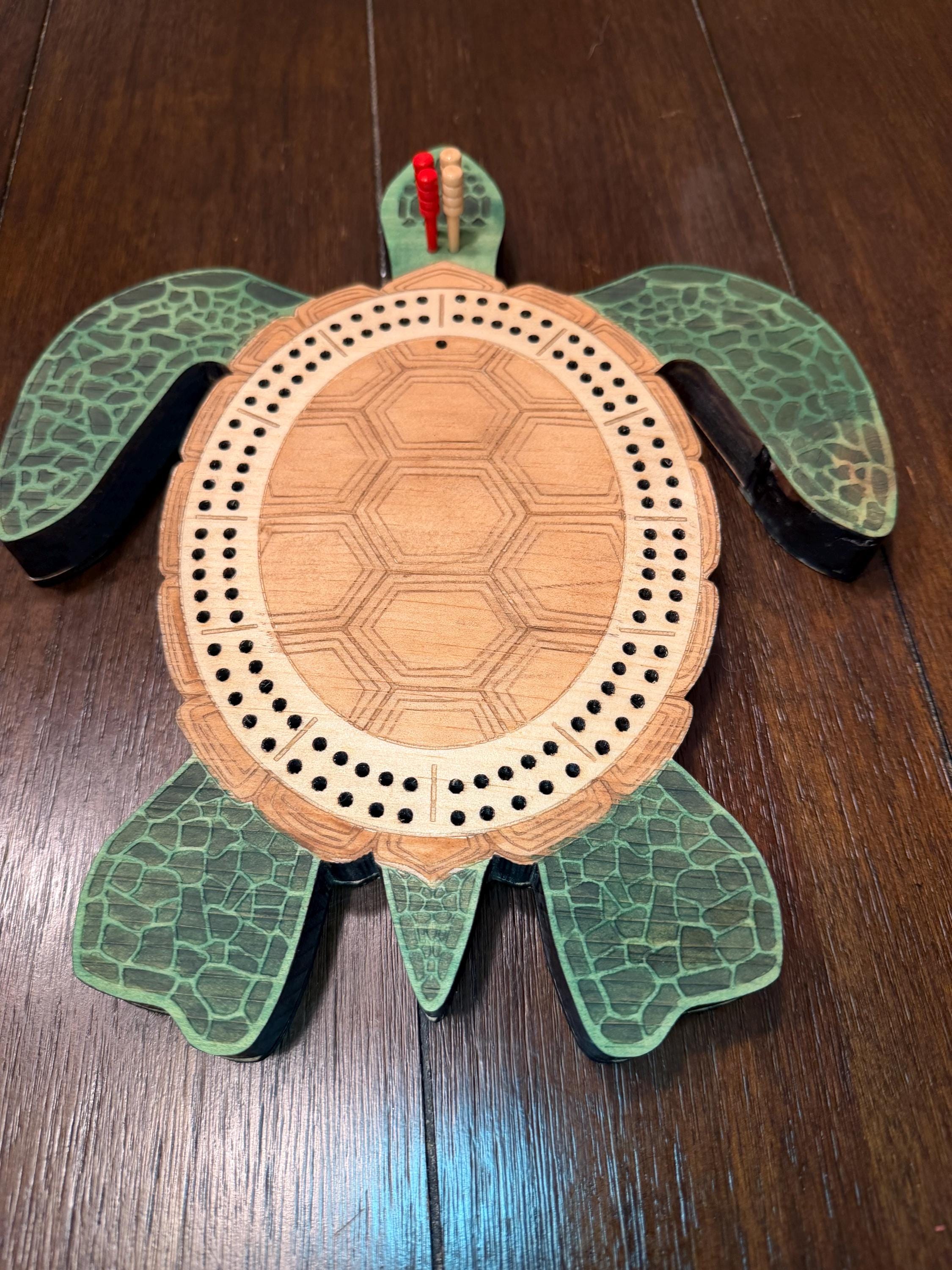 Overhead view of a wooden turtle cribbage board with four cribbage pegs placed at the starting area, showing the full board layout and engraved details.