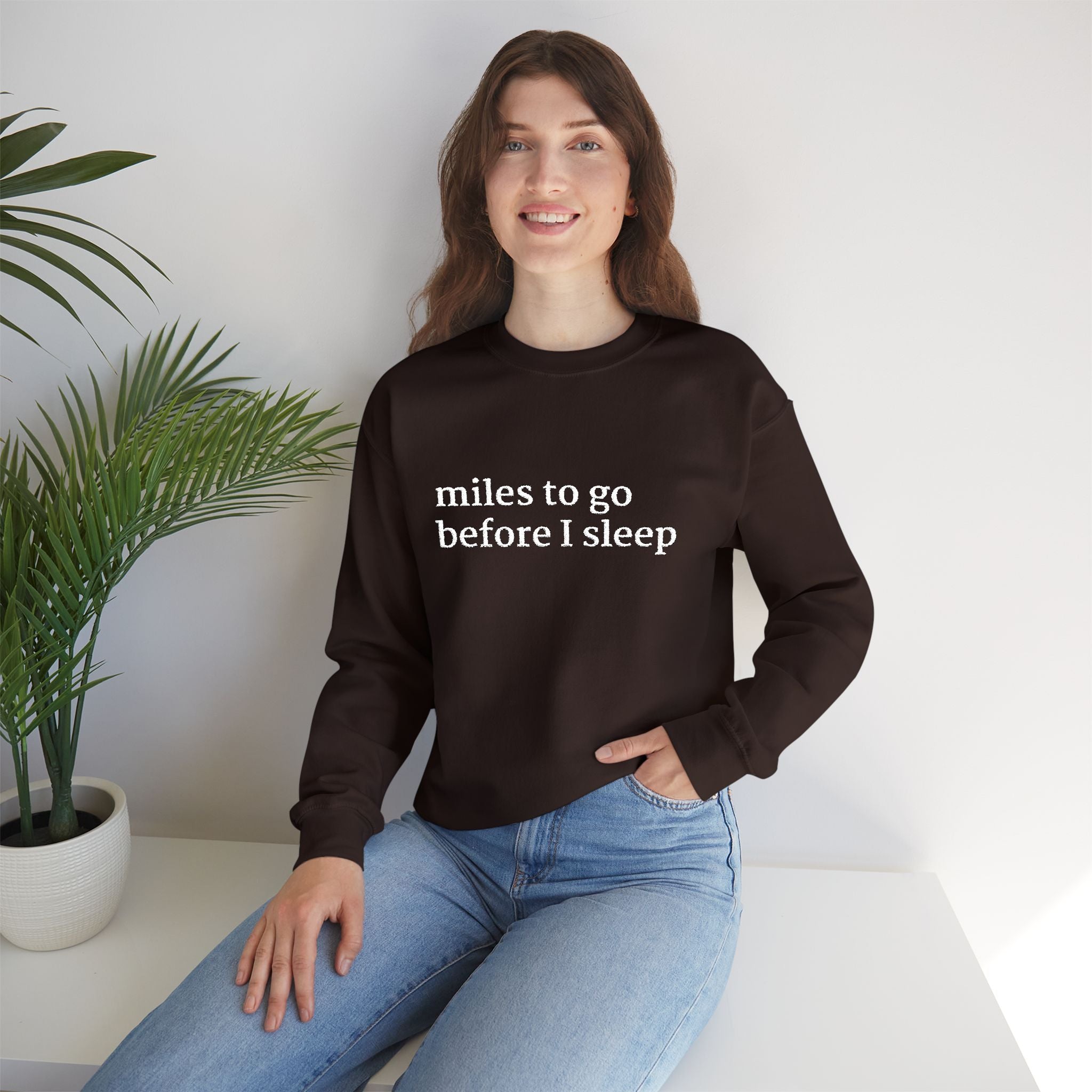 Model wearing a chocolate sweatshirt with 'miles to go before I sleep' text, sitting on a white couch with a plant in the background.