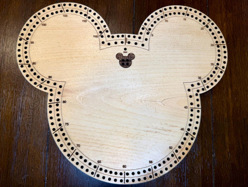Top-down view of a handmade wooden mouse-shaped cribbage board with engraved scoring tracks and numbered holes, photographed on a dark wood surface.
